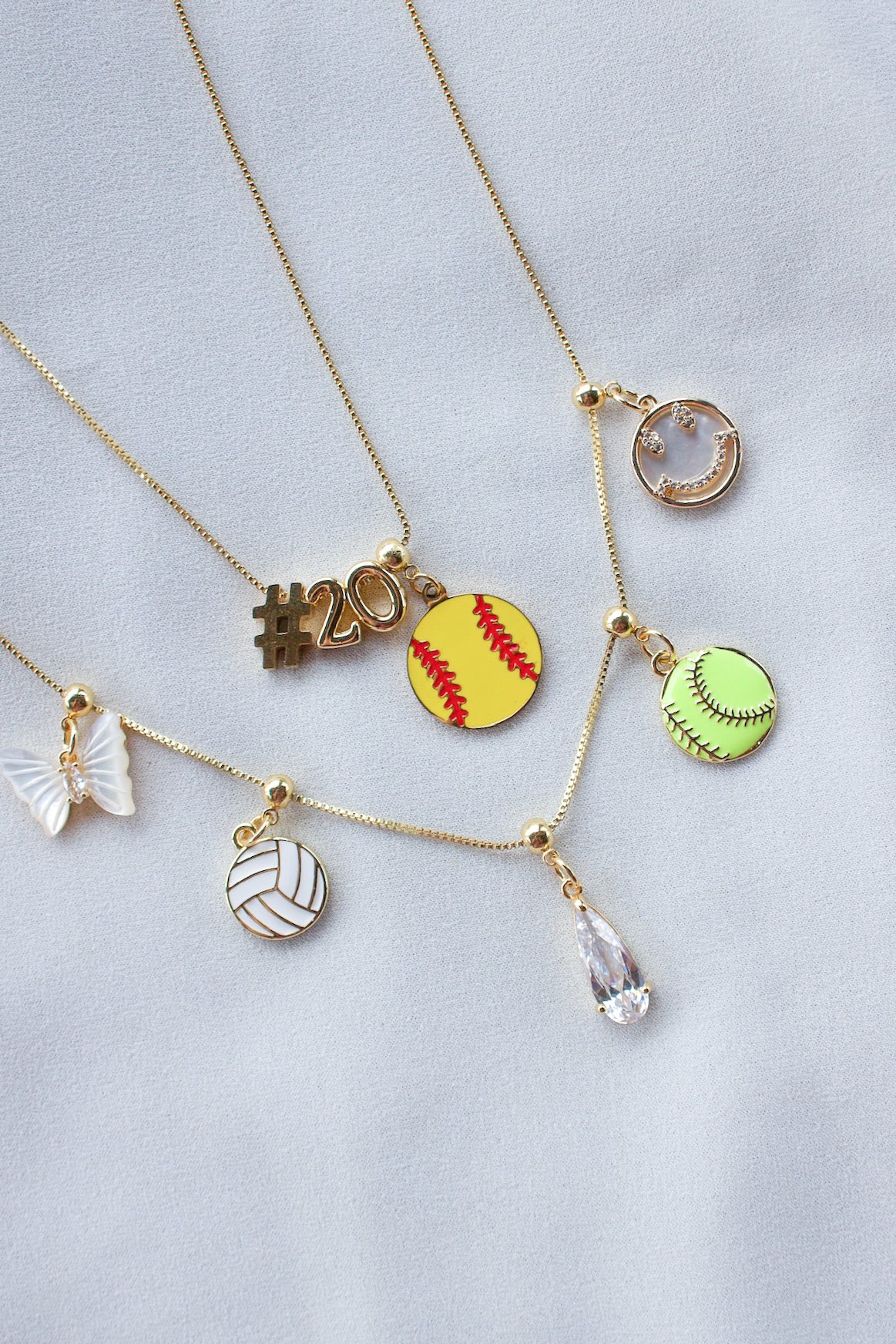 Five Kinsey Designs Butterfly Pearl Grip Charm gold necklaces are displayed on white fabric, featuring butterflies, sports themes, numbers, a crystal drop charm, and a teardrop gem.