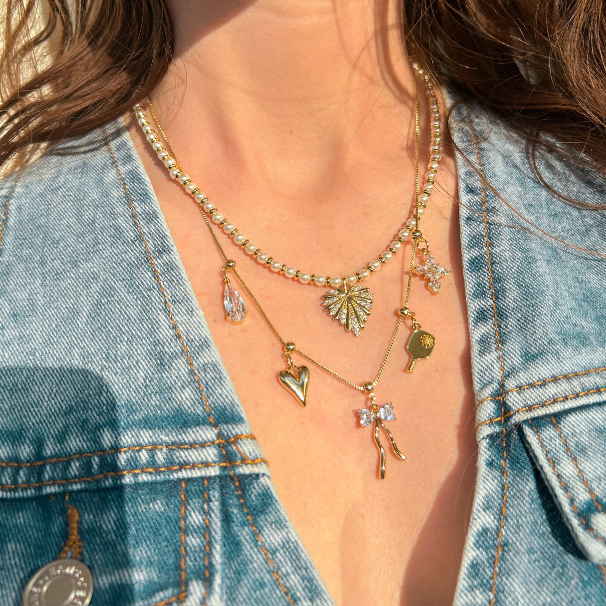 A woman in a denim jacket wears layered gold necklaces from Kinsey Designs, featuring the Gem Bow Grip Charm, as sunlight illuminates her chest.