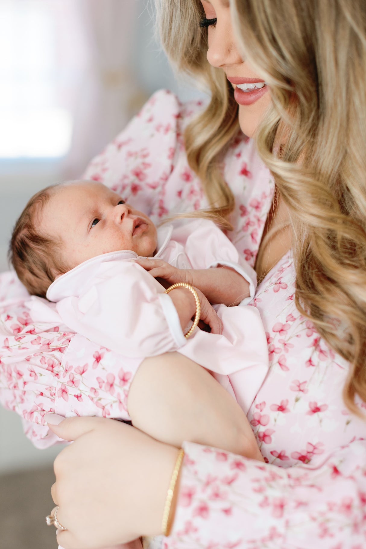 A woman in a floral dress holds her baby, both smiling and wearing Kinsey Designs’ Fallon Baby Cuff—gold-filled, nickel-free, and delicately adjustable for mom and child.