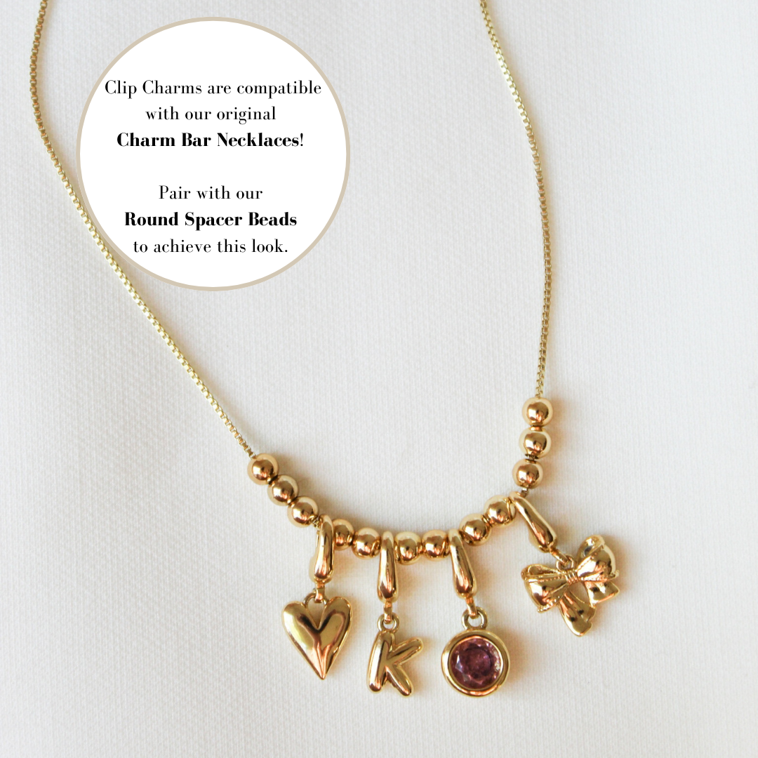 Gold filled charm necklace from Kinsey Designs featuring a heart, letter K, purple gem, bow, and their Baby Bottle Clip Charm, displayed on a white background.