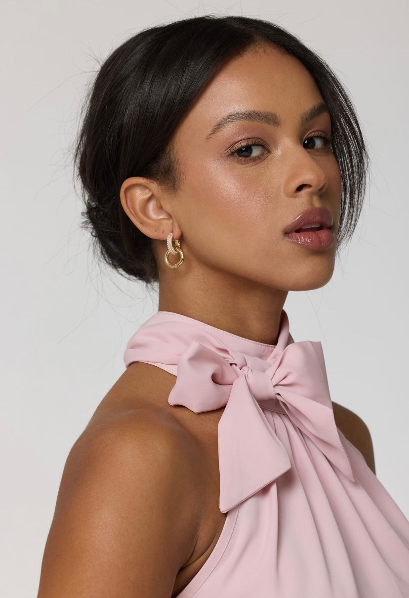 Woman wearing a pink halter top with a bow and natural makeup, looking to the side while accessorizing with Kinsey Designs' Holland Pearl Hoops.