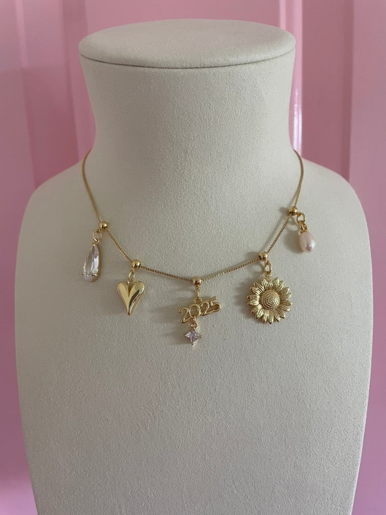 Gold necklace with five charms—teardrop, abstract, 2025 Graduation Grip Charm, sunflower, and pearl—styled on a display stand. Part of Kinsey Designs Archives' Charm Bar Necklaces, perfect for celebrating special milestones.