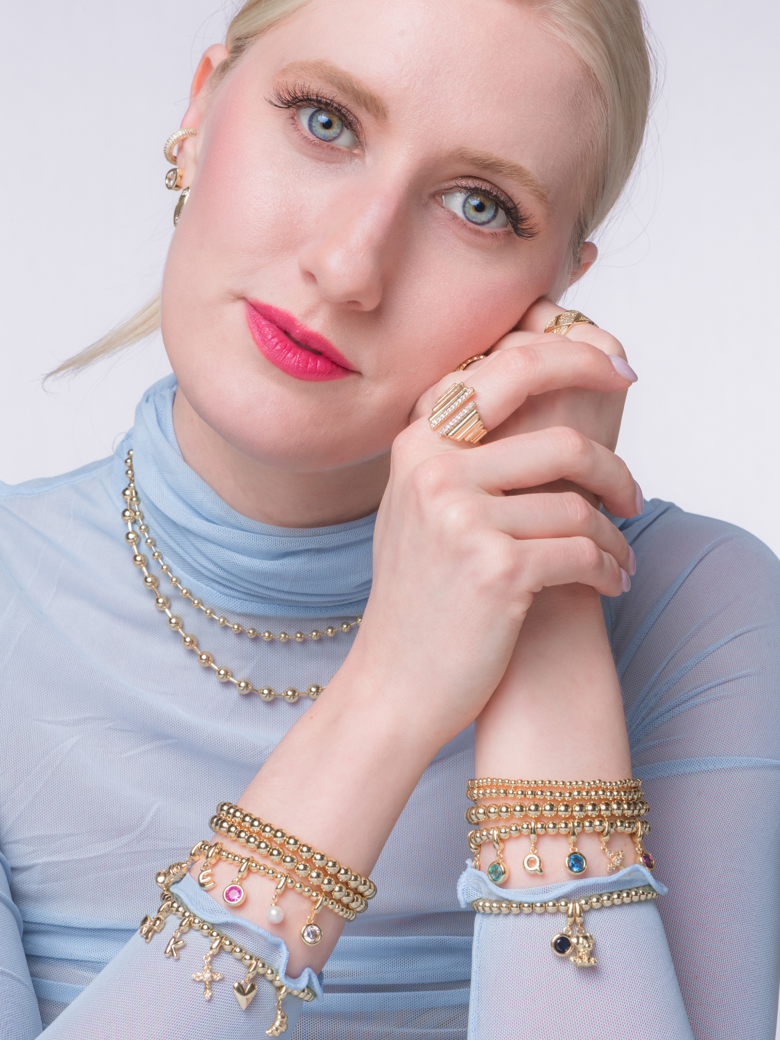 A woman in a light blue top wears gold filled bracelets, rings, and earrings by Kinsey Designs, posing with hands clasped. The delicate Croissant Clip Charm adds a playful touch to her elegant style.