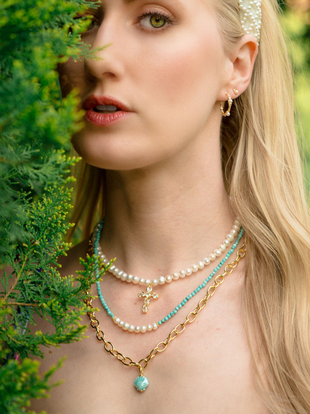 A blonde woman wearing the Arista Shell Pendant Necklace by Kinsey Designs, layered with gold and pearl jewelry, is partially hidden by lush green foliage.