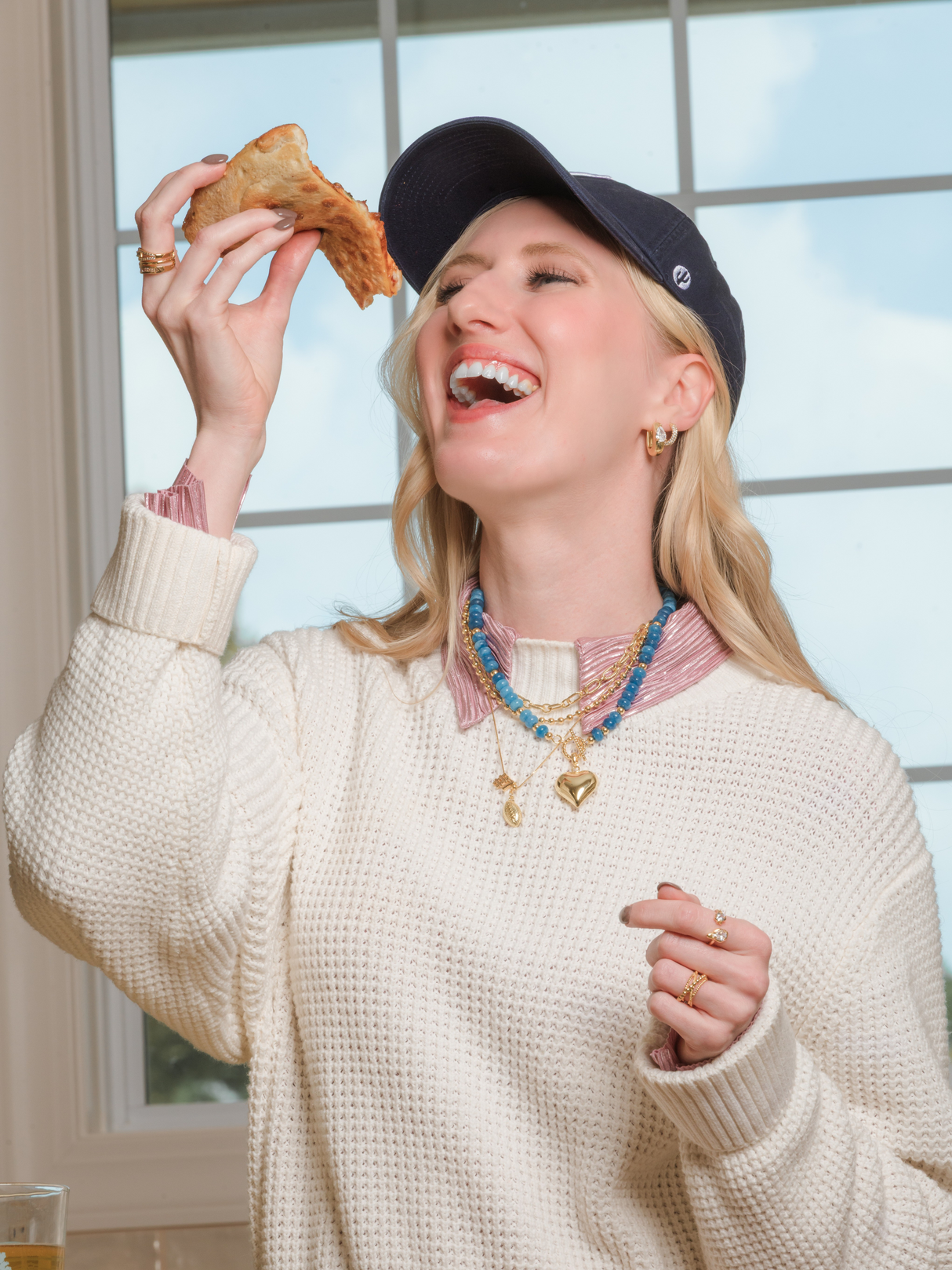 A woman in a sweater and cap laughs indoors, holding pizza near her mouth, her Aspen Puffy Heart Gemstone Necklace by Kinsey Designs adding charm to the cozy moment.