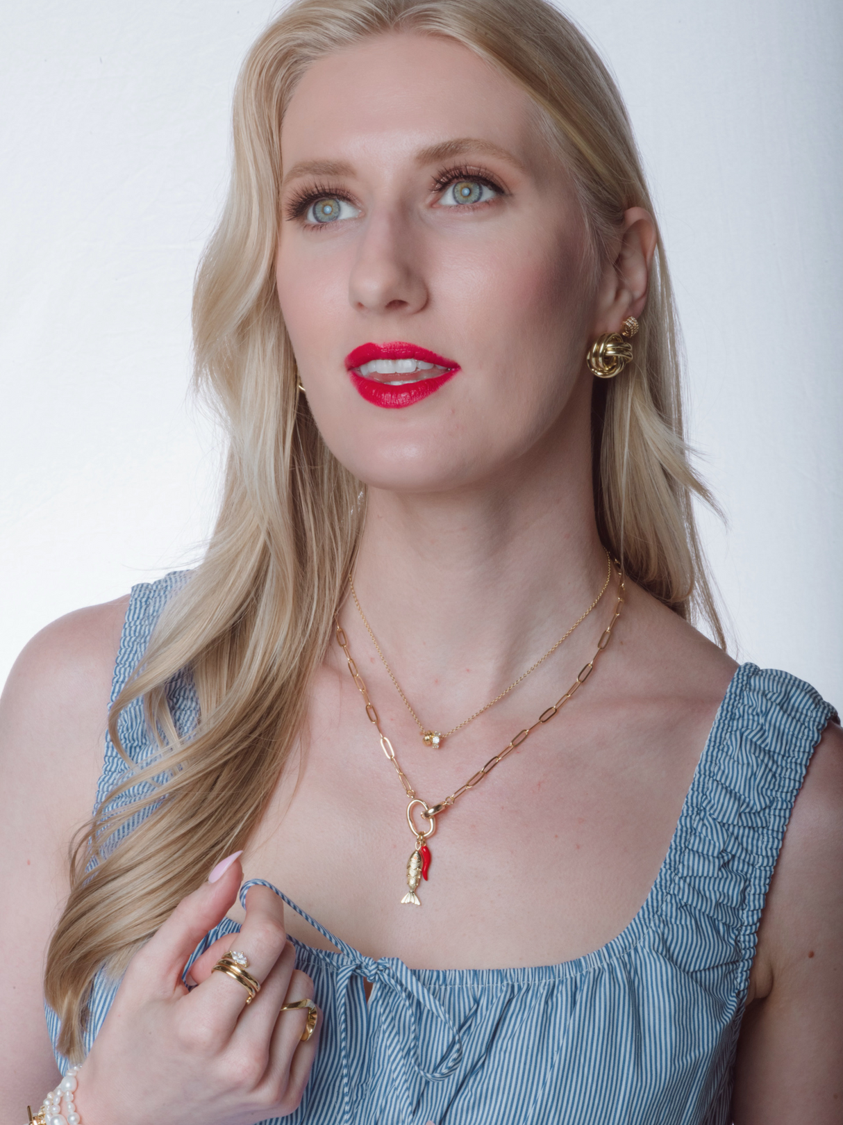 Blonde woman in a blue dress with red lipstick, adorned in gold jewelry including the Kinsey Designs Acqua Pazza Italian Fish Necklace, looking up against a white background.