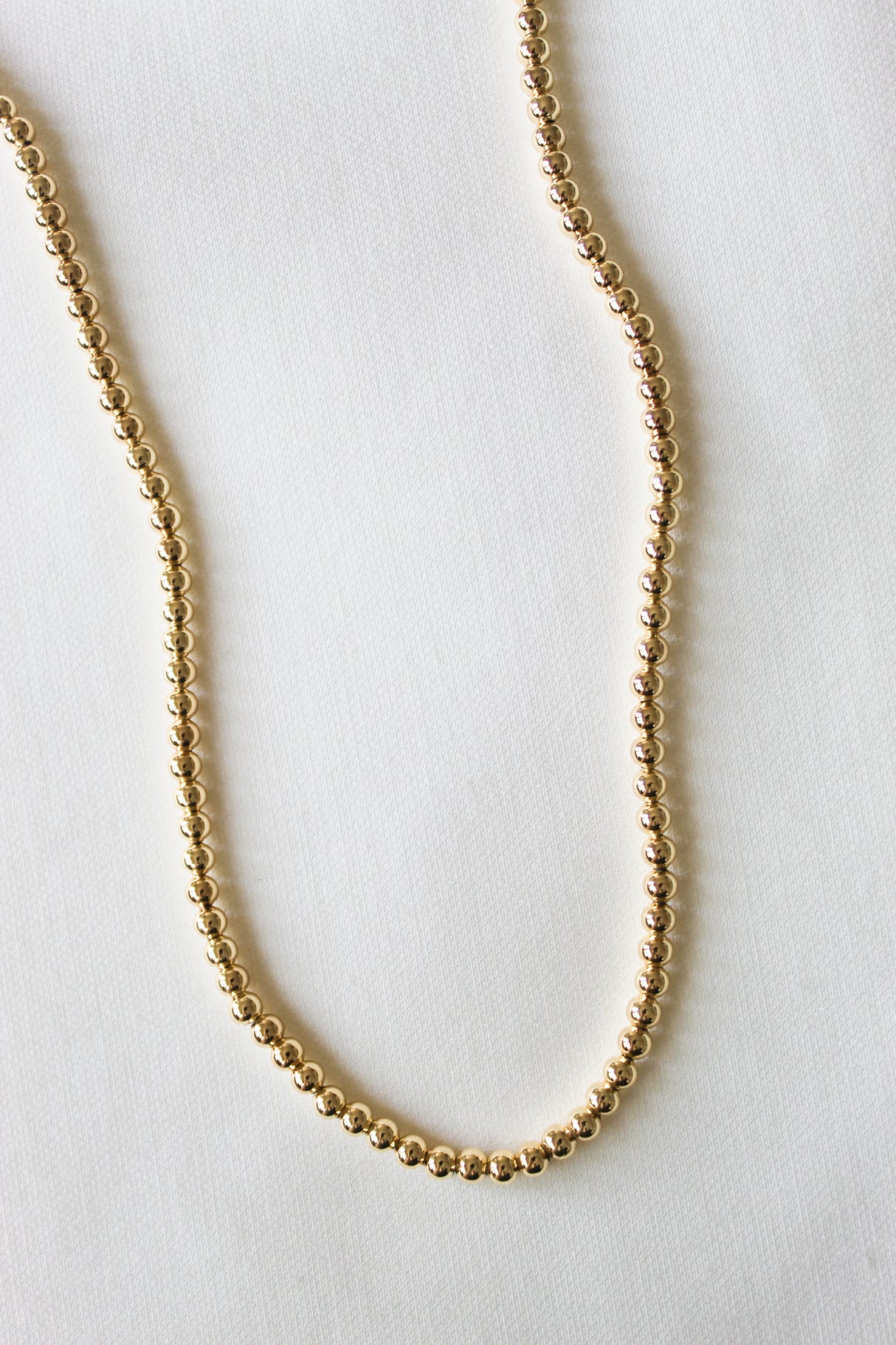 The Adda Necklace 3mm by Kinsey Designs is a minimalist gold beaded necklace displayed in a soft curve on a white textured surface, perfect for those who appreciate simple, elegant jewelry.