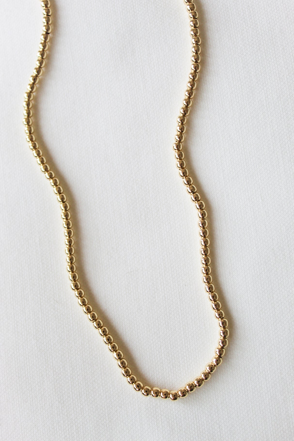 The Kinsey Designs Adda Necklace 4mm, a gold beaded piece displayed in a gentle curve on a white textured surface, is ideal for lovers of minimalist jewelry.
