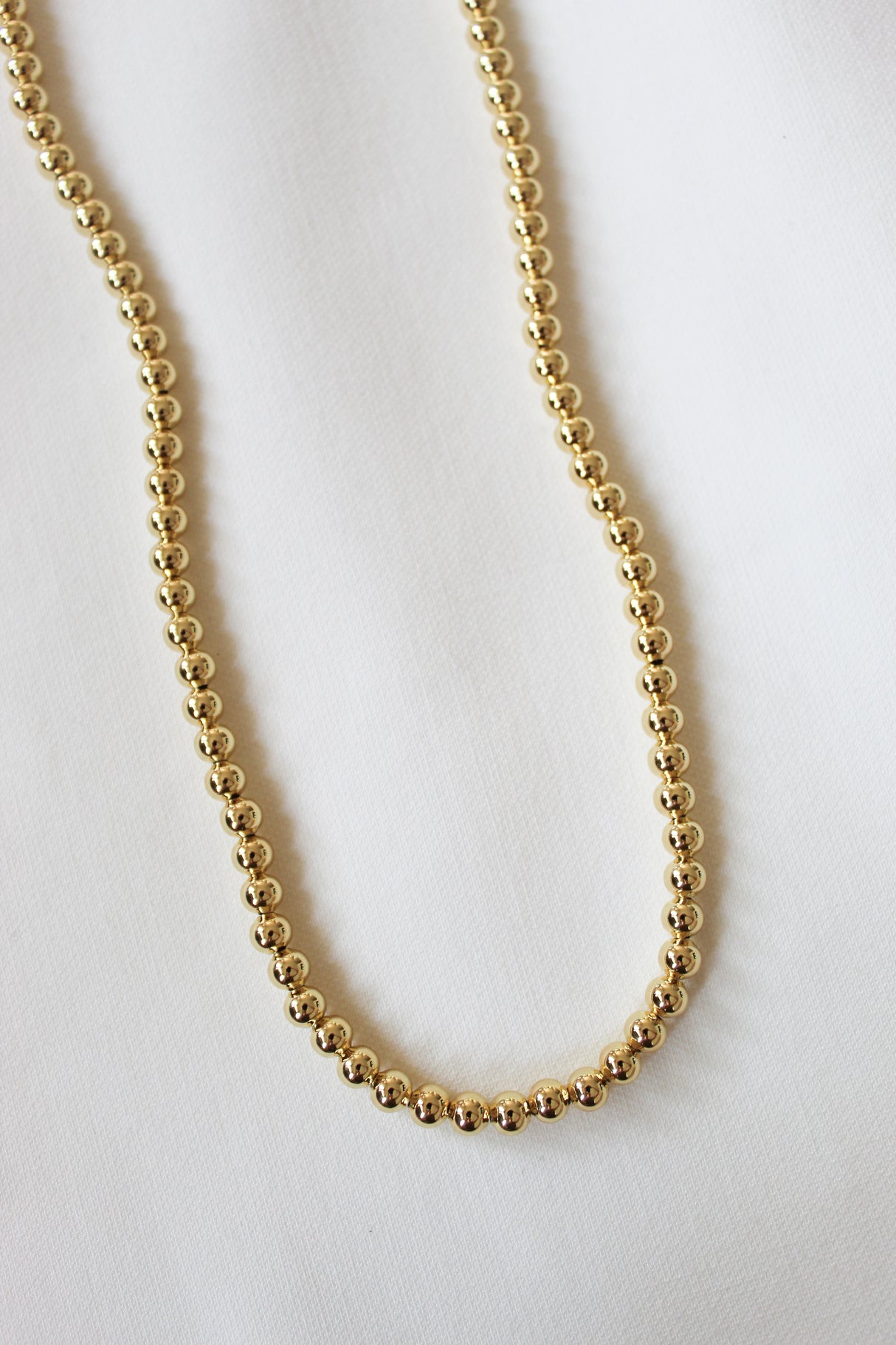 The Kinsey Designs Adda Necklace 5mm, a gold beaded piece from the Adda collection, is shown on white fabric—ideal for minimalist jewelry lovers.