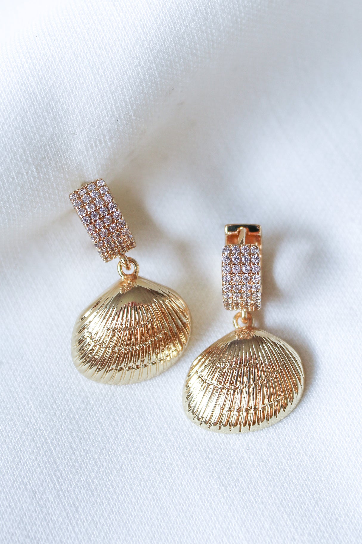 Discover the Alana Huggies by Kinsey Designs Archives—gold-filled shell-shaped earrings with textured surfaces and sparkling encrusted tops, featured in our sample sale and displayed on a white fabric background.