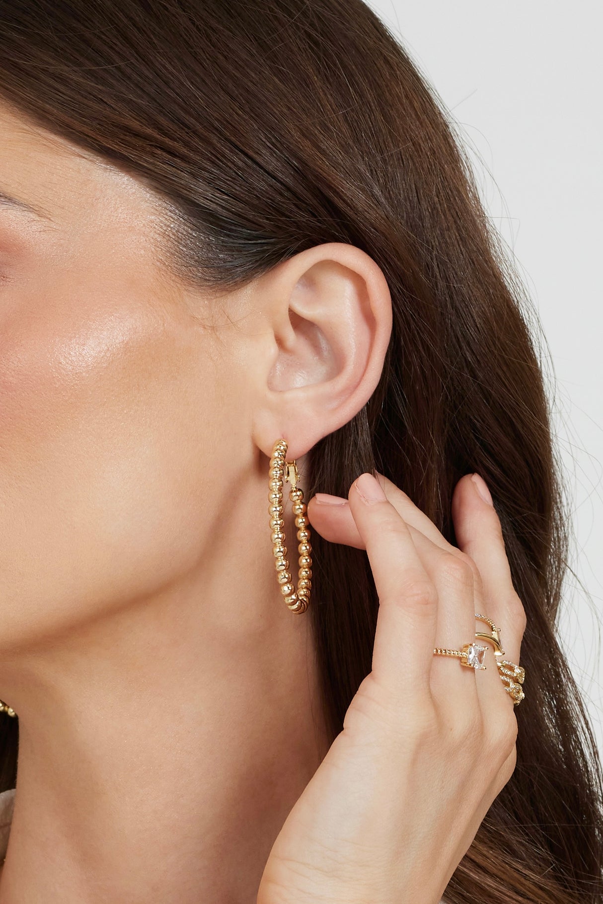 A woman touches her ear with manicured fingers, showcasing Kinsey Designs Archives’ Atlas Hoop earrings and gold rings for an effortlessly chic sample sale look.