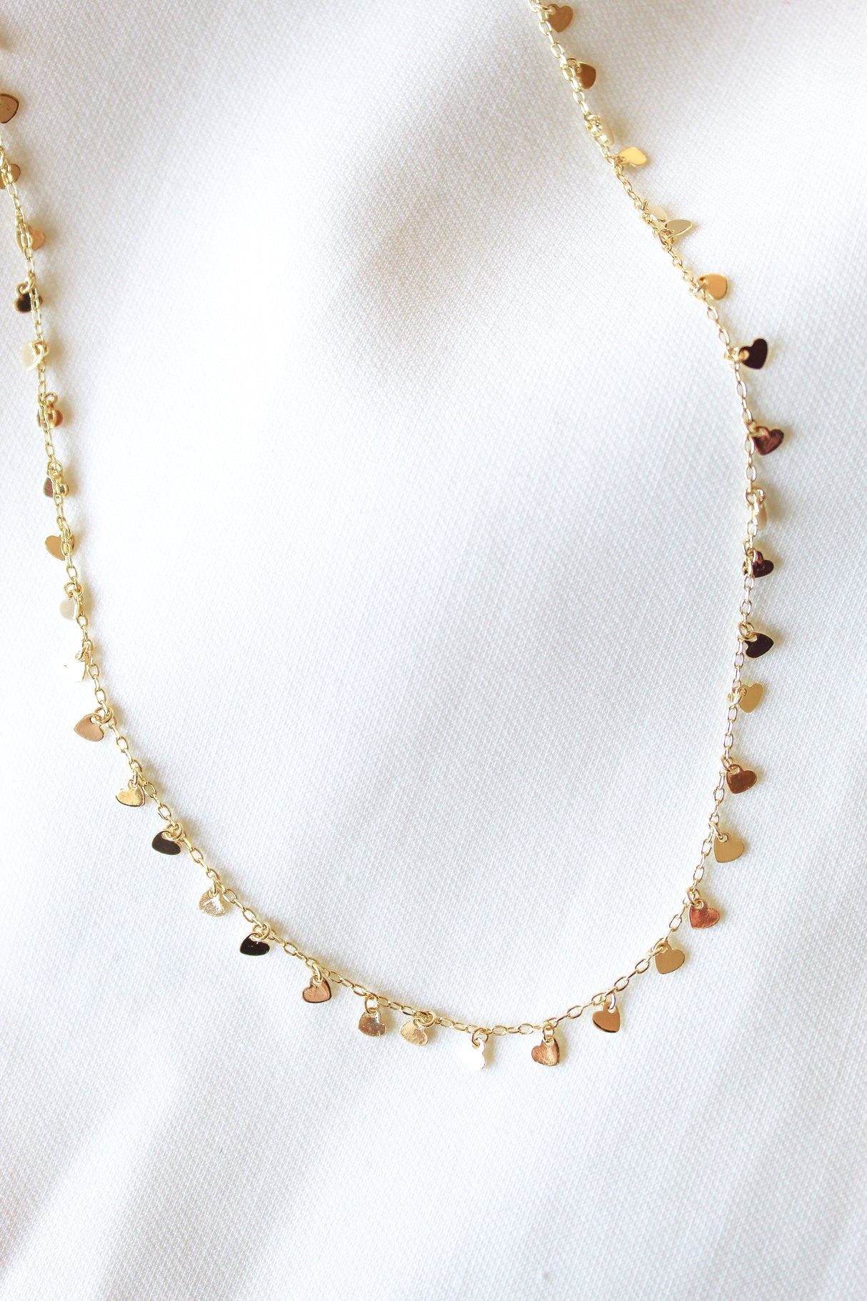 The Avery Necklace by Kinsey Designs Archives features gold-filled small heart-shaped charms on a white textured fabric background. This 14-inch necklace includes an extender for an adjustable, perfect fit.