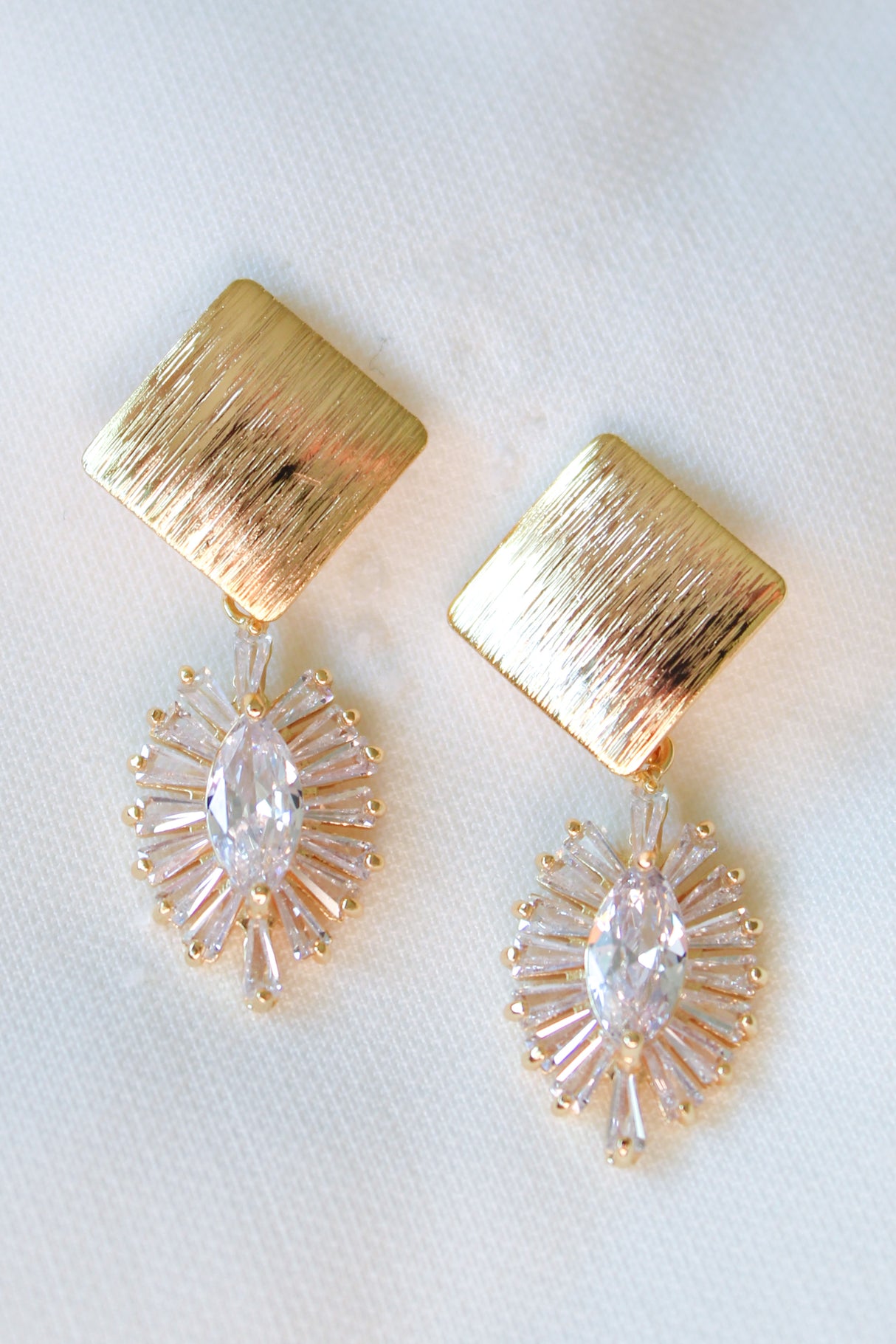 The Aza Earrings by Kinsey Designs Archives feature gold-filled, textured square tops and oval crystal drops. These elegant 1.5-inch earrings are showcased beautifully on a white fabric background.