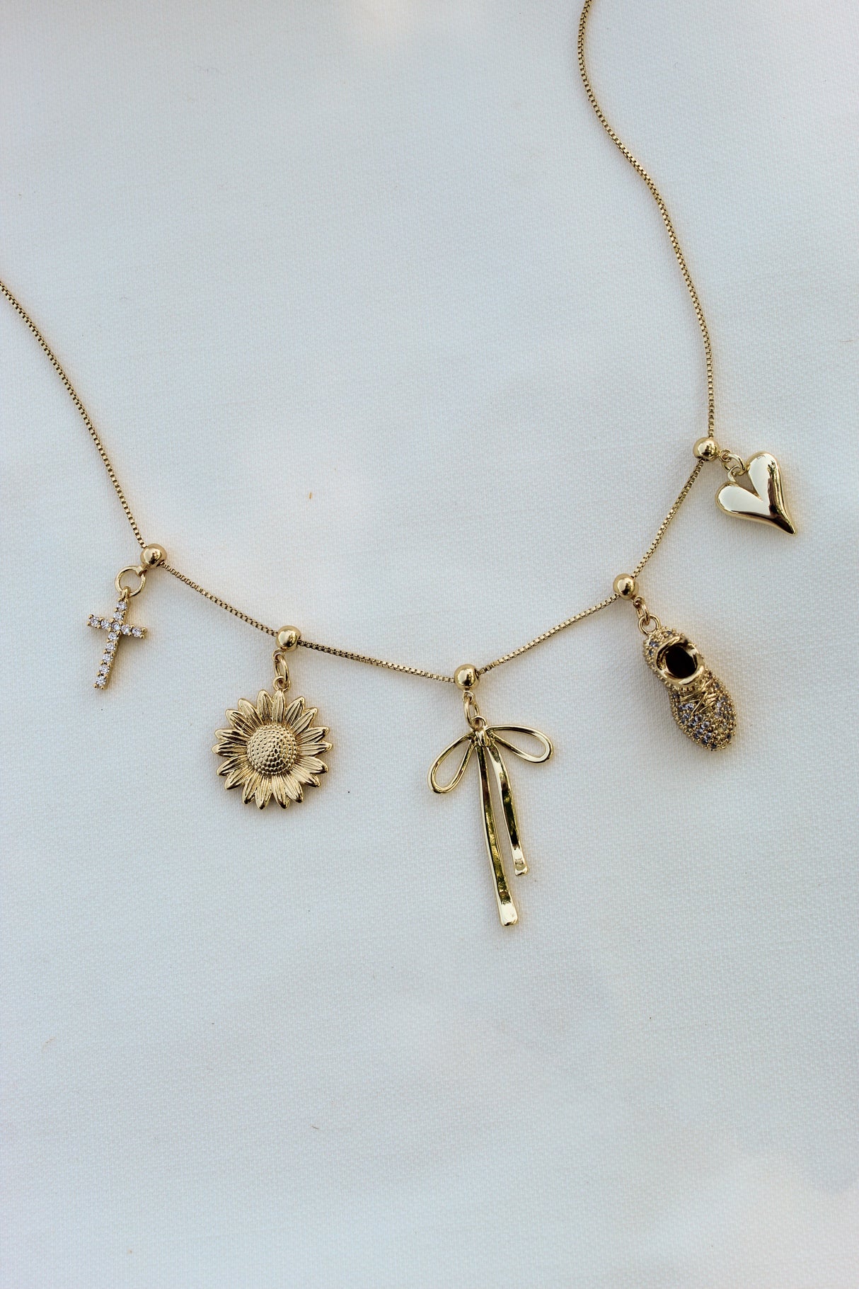 The Sunflower Grip Charm by Kinsey Designs is a gold bar necklace featuring a sunflower charm with cross, bow, jeweled boot, and heart accents—an elegant nature-inspired jewelry piece shown on a white background.