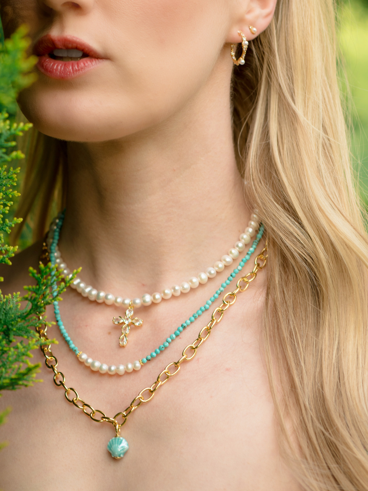 A woman with long blonde hair and a hoop earring wears the Bellena Pearl Cross Necklace by Kinsey Designs—layered gold, pearl, and turquoise jewelry featuring a crystal-studded cross pendant.