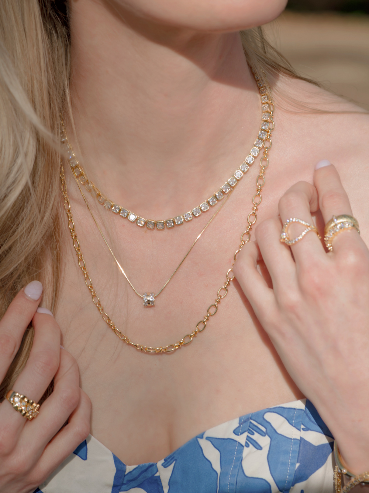 A woman in a blue and white floral top adjusts her gold jewelry, featuring the Kinsey Designs Blaire Square Tennis Necklace and rings.