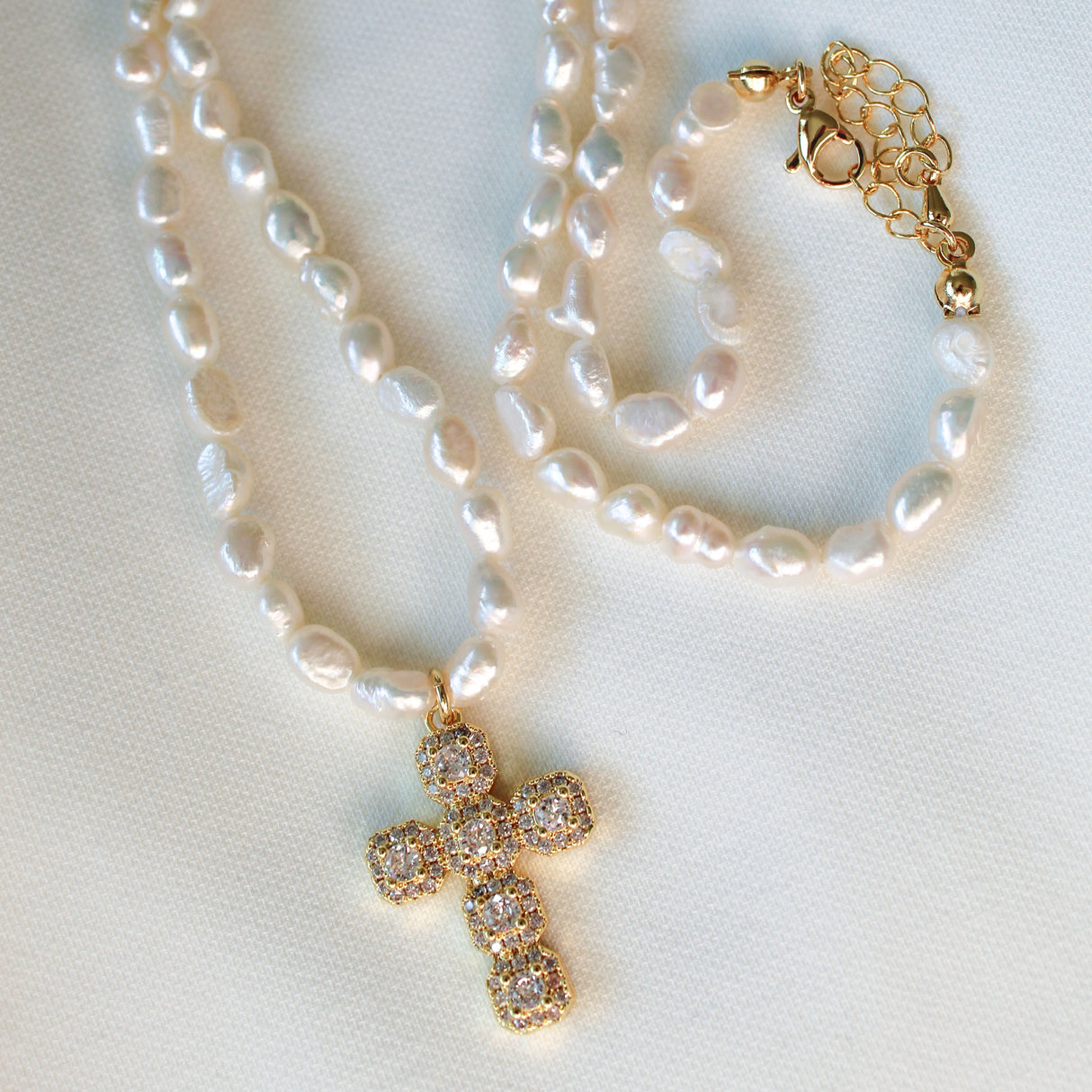 The Baldwin Pearl Necklace by Kinsey Designs features lustrous freshwater pearls and a gold-filled cross pendant with diamond accents, shown on a white fabric background.