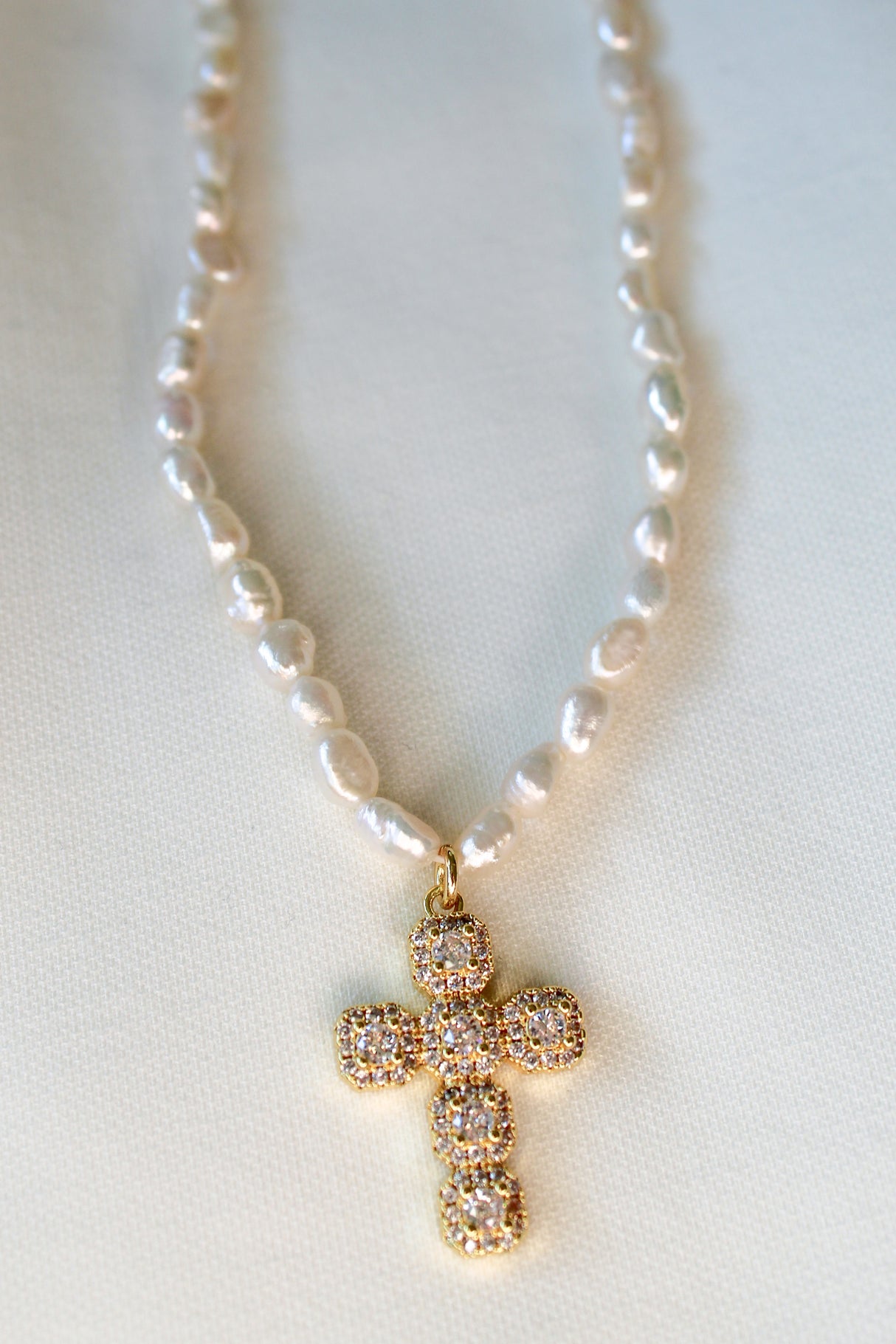 The Baldwin Pearl Necklace by Kinsey Designs features lustrous freshwater pearls and a gold-filled cross pendant with diamonds, elegantly displayed on a light fabric background.