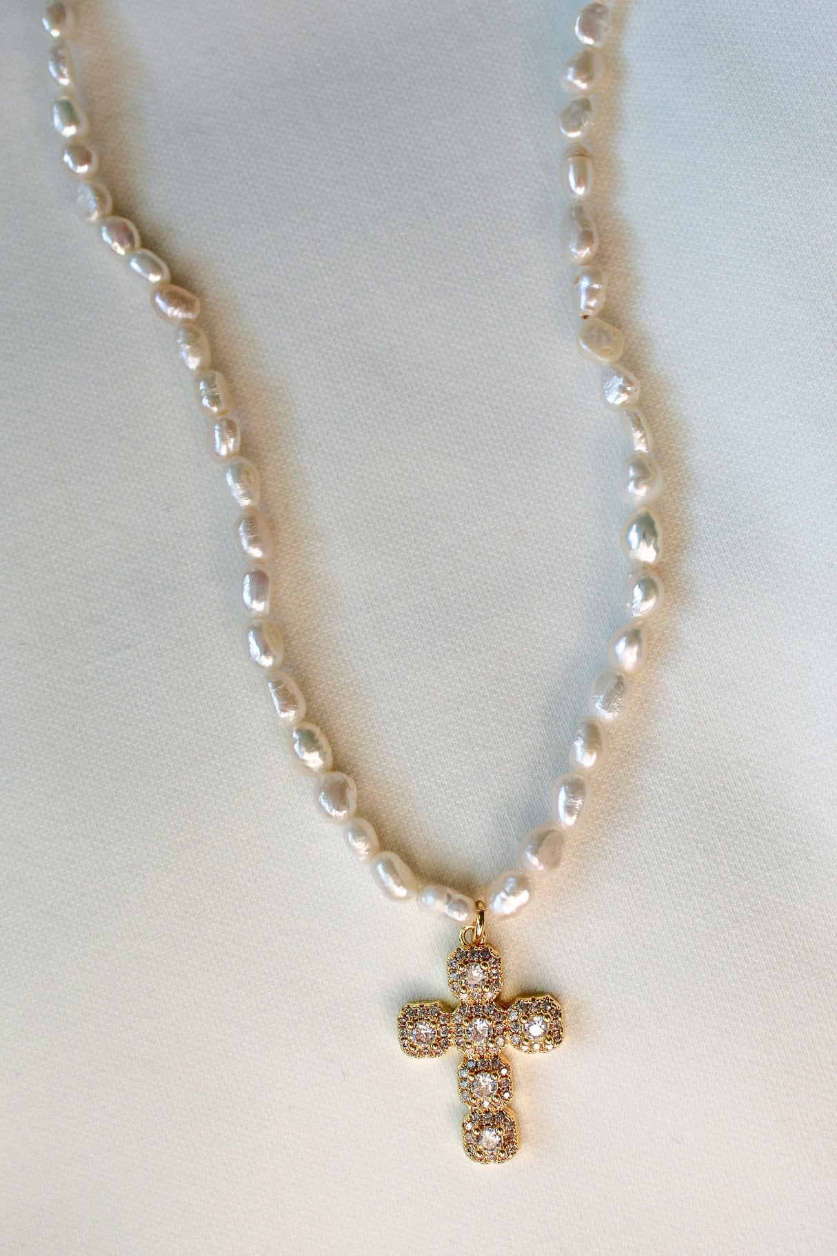 The Baldwin Pearl Necklace by Kinsey Designs features freshwater pearls and a jeweled gold-filled cross pendant, elegantly displayed on a white fabric background.