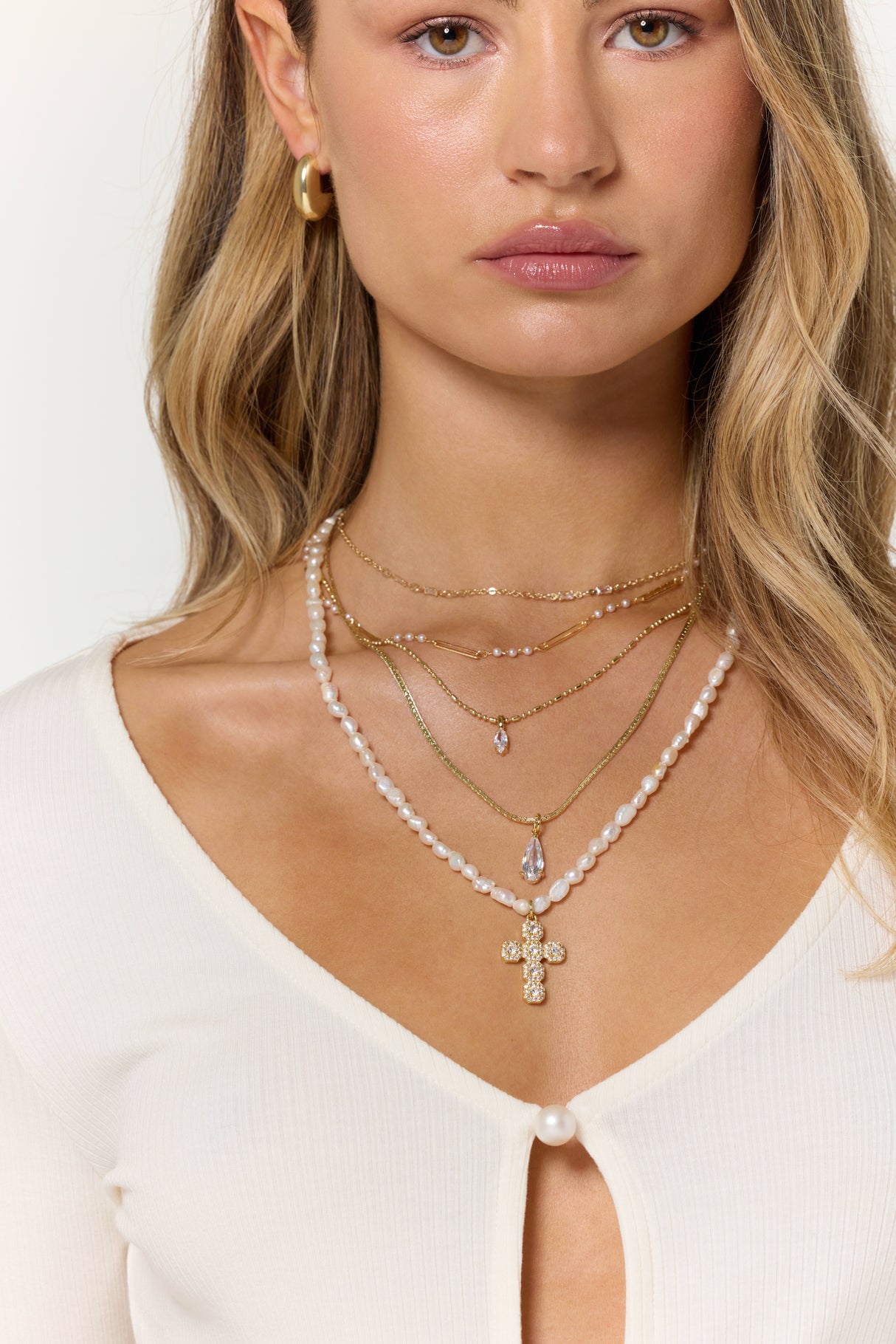 A woman in a white top and gold hoop earring wears layered necklaces, featuring Kinsey Designs' Baldwin Pearl Necklace with freshwater pearls and a gold filled cross pendant.