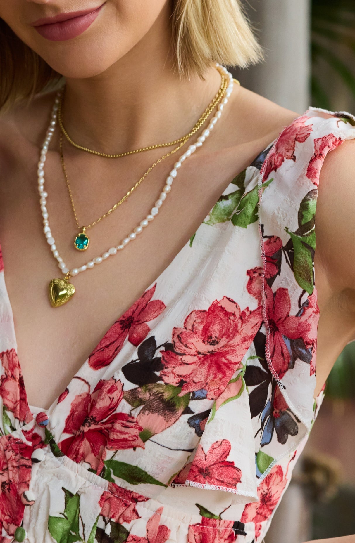 A woman wears layered necklaces, including a gold heart locket and the Kinsey Designs Giselle Pearl Necklace, strung with luminous freshwater pearls, paired with a floral dress.