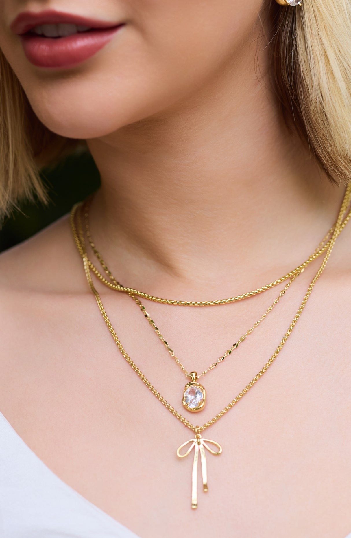 A woman wears layered gold-filled necklaces from Kinsey Designs, including the Birdie Necklace with a bow charm and another necklace with a cushion-cut CZ stone pendant.