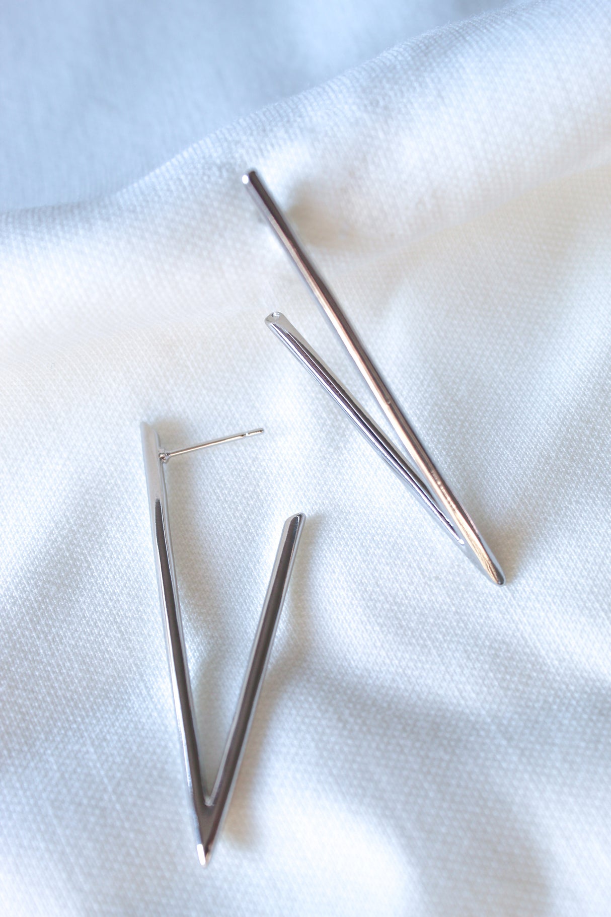 The Bowen Earrings by Kinsey Designs are silver, V-shaped, rhodium-plated earrings shown on a white textured fabric background.