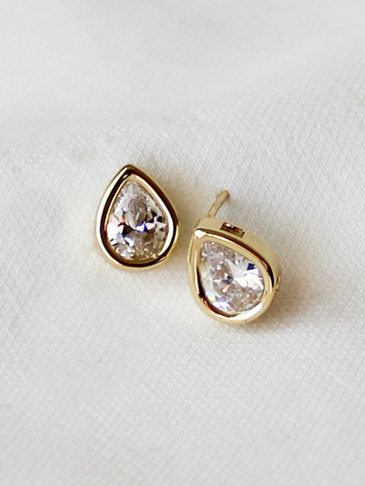 The Bridget Stud Earrings by Kinsey Designs feature pear-shaped clear gemstones set in gold fill, displayed on a white fabric background—perfect for adding elegant style to your jewelry collection.