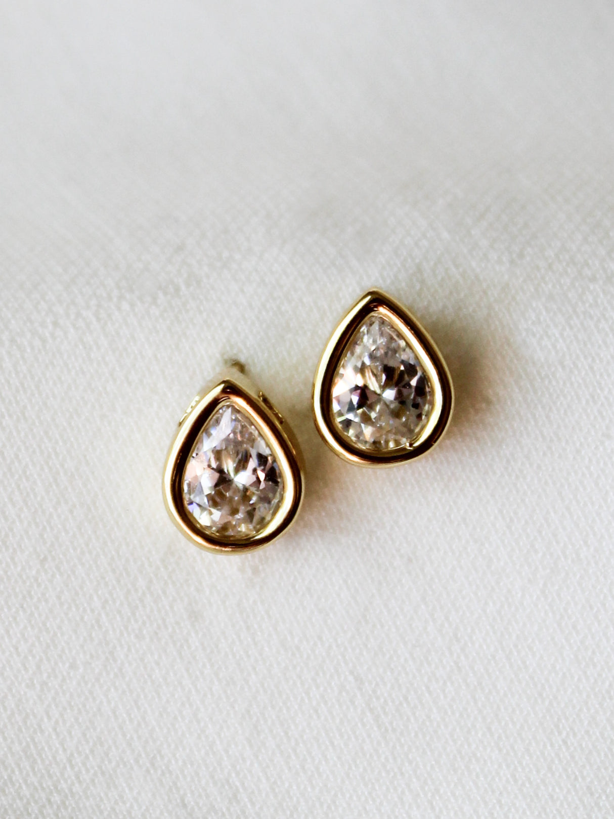 The Bridget Stud Earrings by Kinsey Designs feature teardrop-shaped clear gemstones set in gold, elegantly displayed on a white fabric background—an enduringly stylish piece for your jewelry collection.