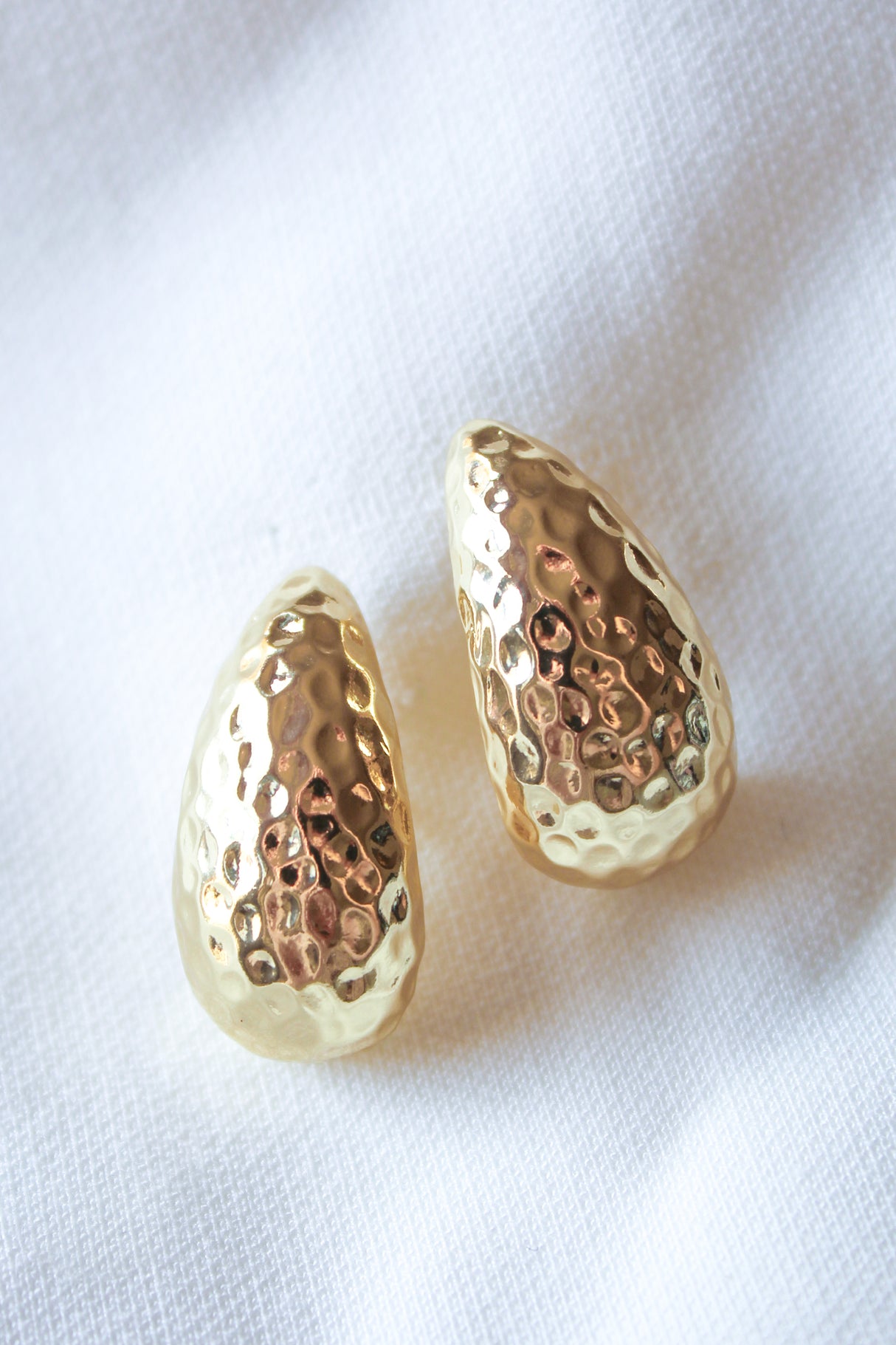 The Surtur Earrings by Kinsey Designs Archives, each 1 inch long with a hammered gold filled finish, are displayed elegantly on a white textured fabric background.