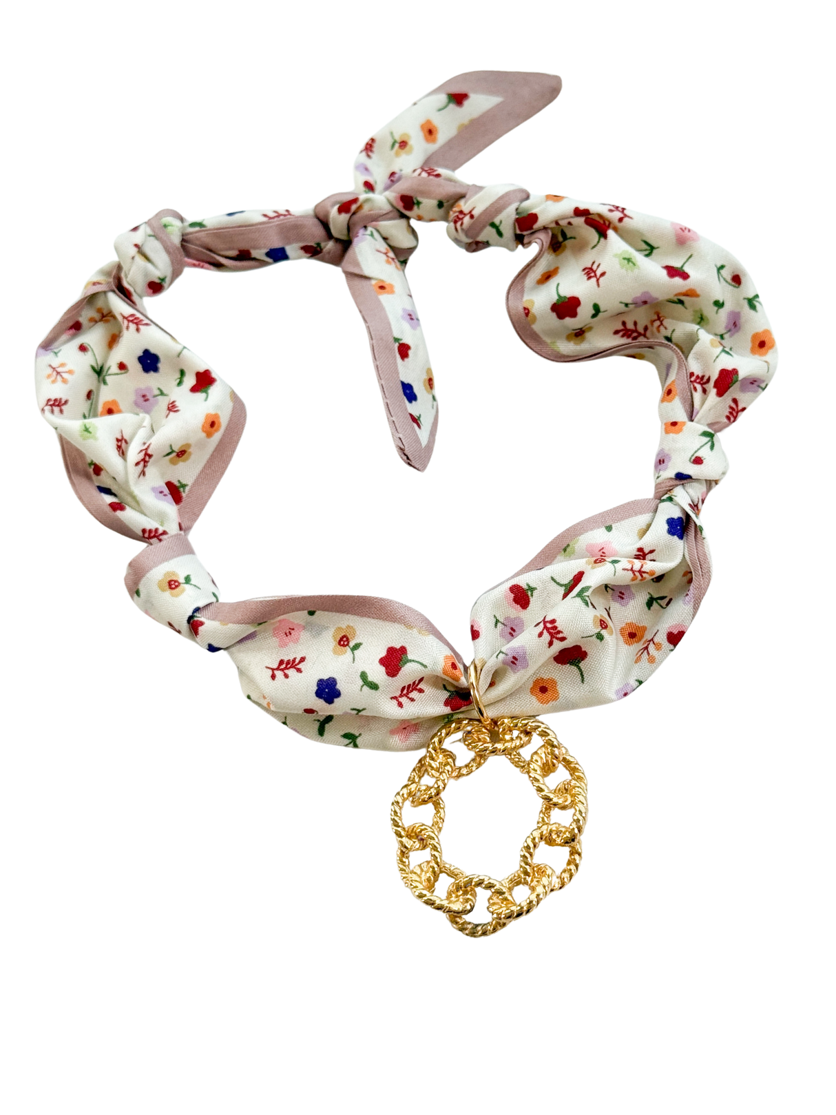 The Calabria Scarf Necklace by Kinsey Designs features a floral fabric with an adjustable design, knotted details, and a large gold chain-link pendant at the center.