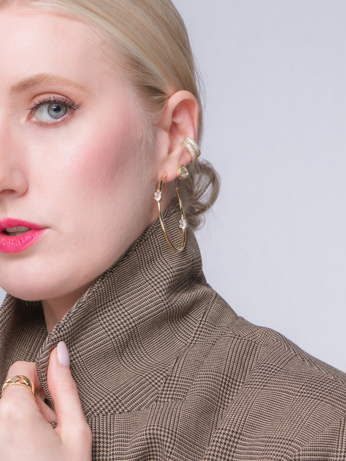 A woman in a plaid blazer wears Kinsey Designs Archives' Clotho Hoop Earrings, along with gold-filled rings, looking to the side against a plain background.