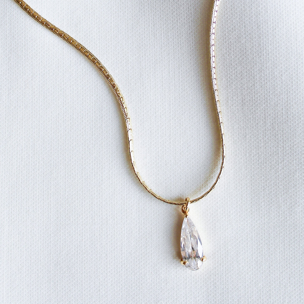 The Charlotte Drop Necklace by Kinsey Designs features a gold filled teardrop design with a clear cubic zirconia pendant, elegantly showcased on a white fabric background.