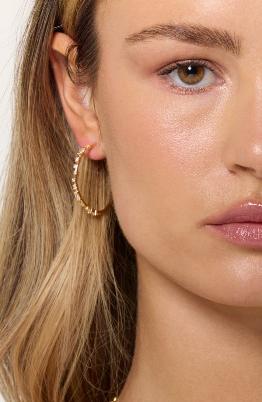Close-up of a woman wearing Kinsey Designs' Chase Hoop Large gold-filled earrings with textured details, highlighting part of her hair and lips.