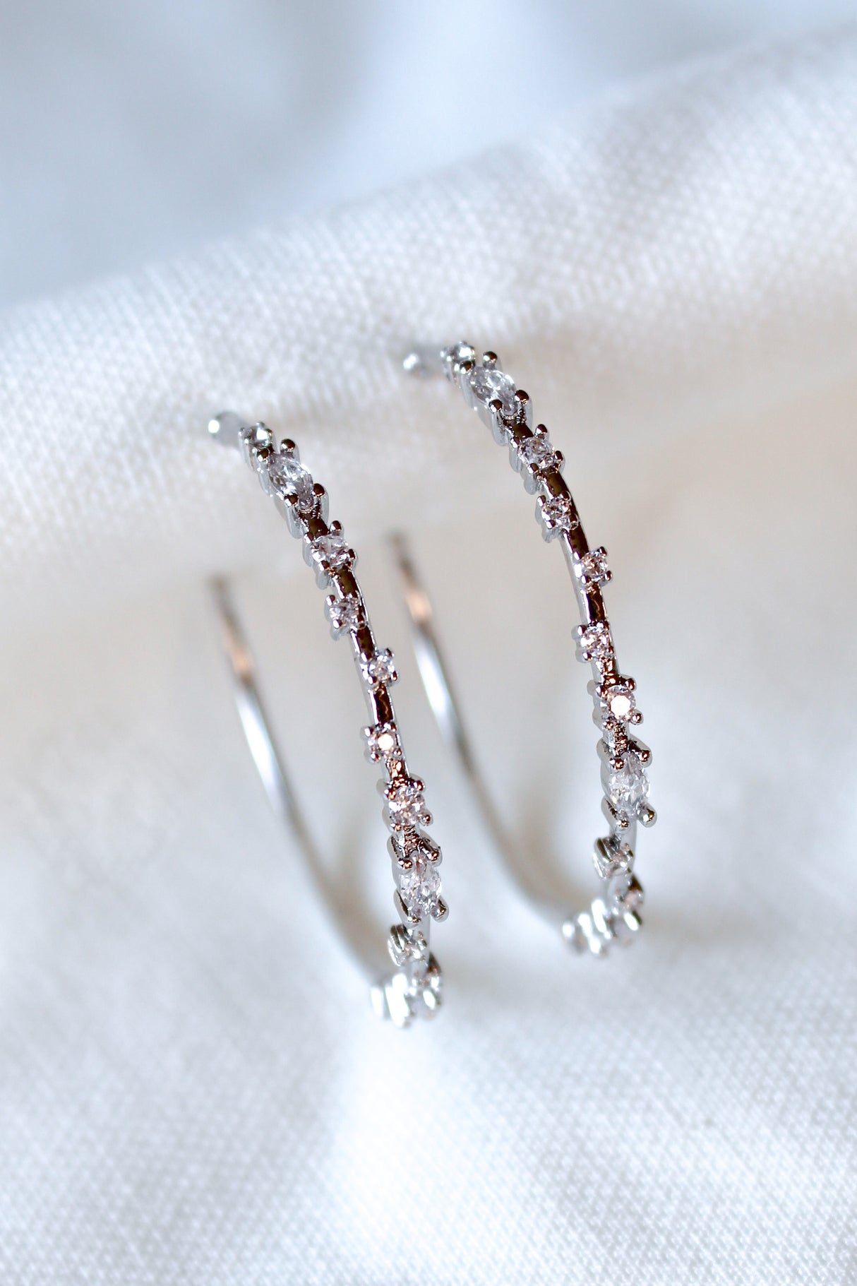 The Chase Hoop Large earrings by Kinsey Designs are silver hoops embellished with sparkling cubic zirconia, shown on a white fabric background.