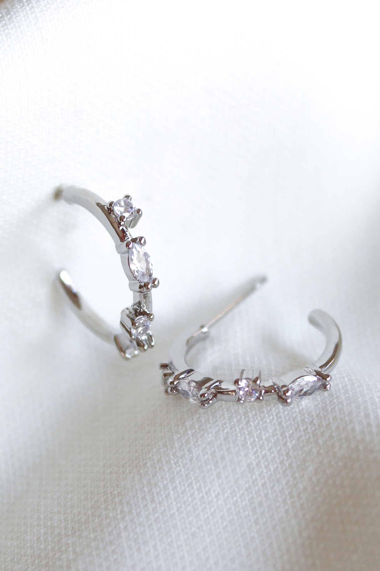 The Kinsey Designs Chase Hoop Mini Silver earrings feature two 0.5" diameter silver hoops adorned with small clear cubic zirconia gemstones, shown resting on a white textured fabric.