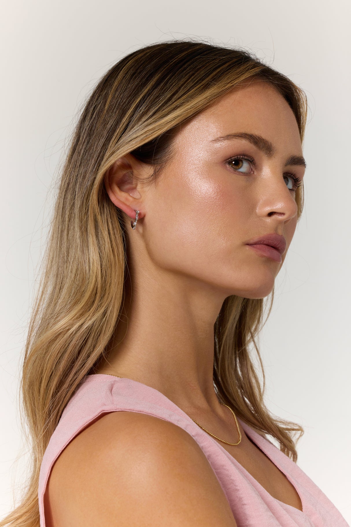 A woman with long, light brown hair wearing a pink top, gold necklace, and Kinsey Designs’ Chase Hoop Mini Silver earrings (.5" diameter), crafted from rhodium plated brass with cubic zirconia accents, looking sideways.