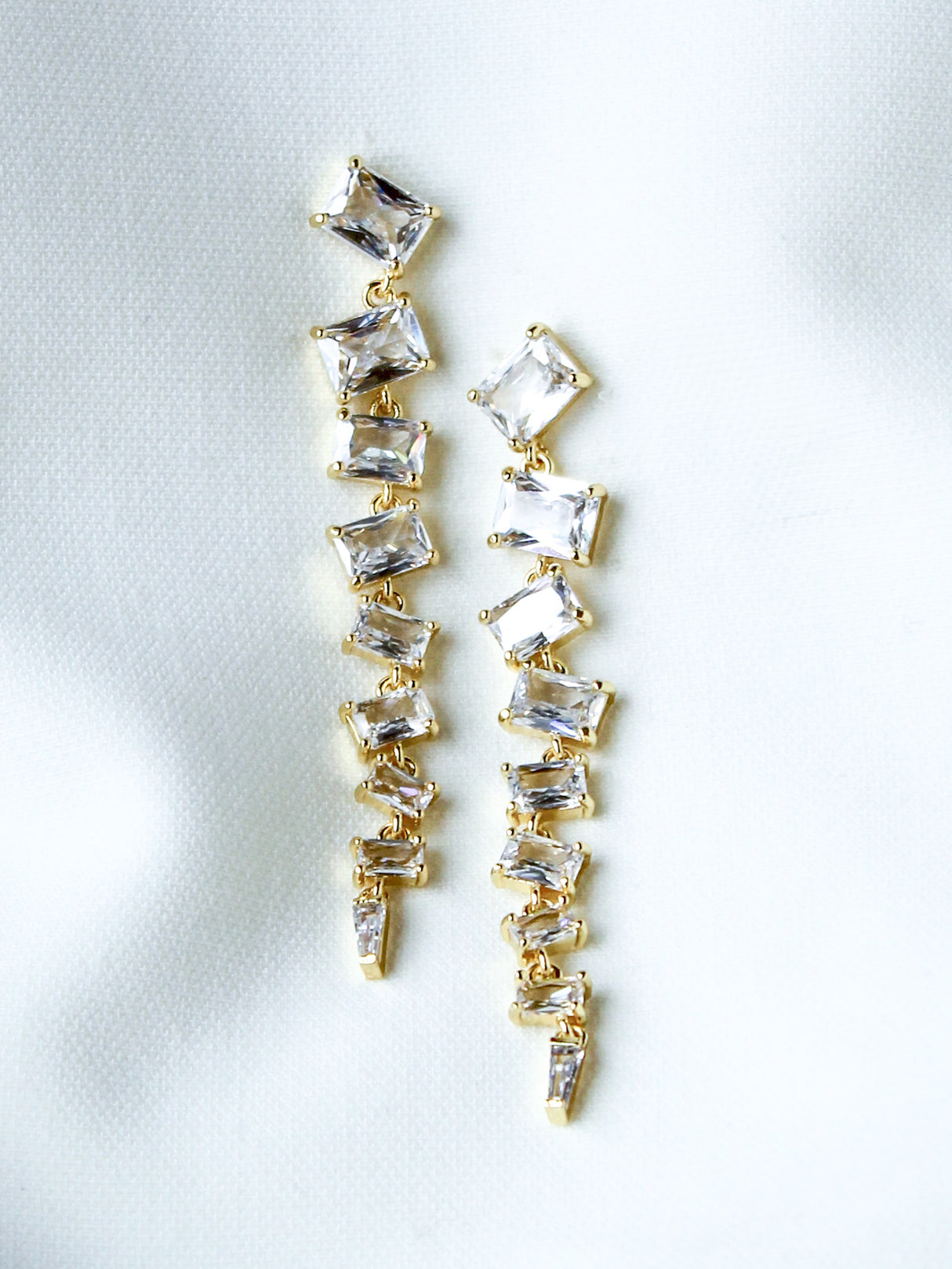 The Cher Drop Earrings by Kinsey Designs are 2.5-inch gold-filled pieces with multiple square and rectangular clear gemstones, shown on white fabric—an elegant choice at a sample sale price.