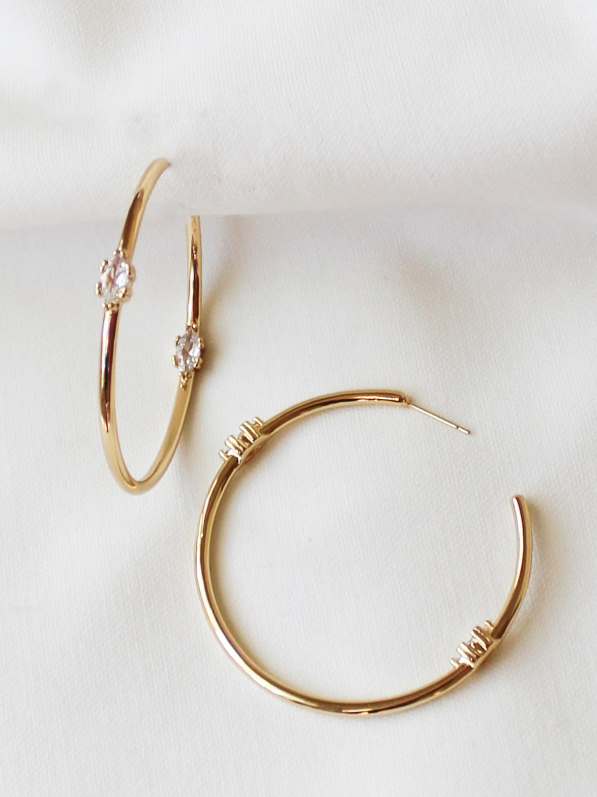 The Clotho Hoop Earrings by Kinsey Designs Archives feature gold-filled hoops with small clear gemstones, resting on white fabric. Part of our exclusive sample sale, they offer a chic look.