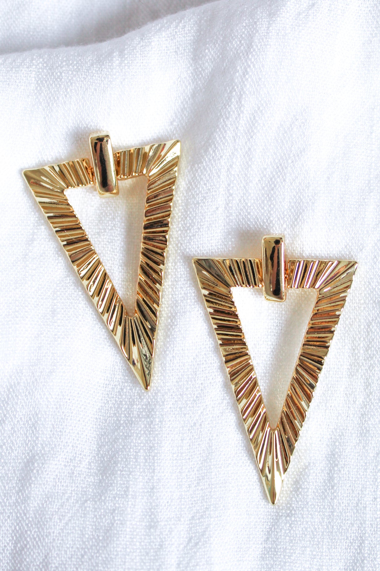 The Colin Earring by Kinsey Designs, featuring a gold textured triangular design, is showcased on a white fabric background.