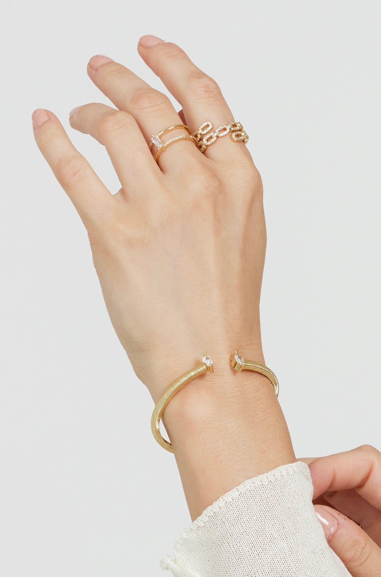 A hand wears Kinsey Designs' Cora Ring—an open gold ring with a baguette CZ stone—and a gold cuff bracelet with crystal ends, set against a plain light background.