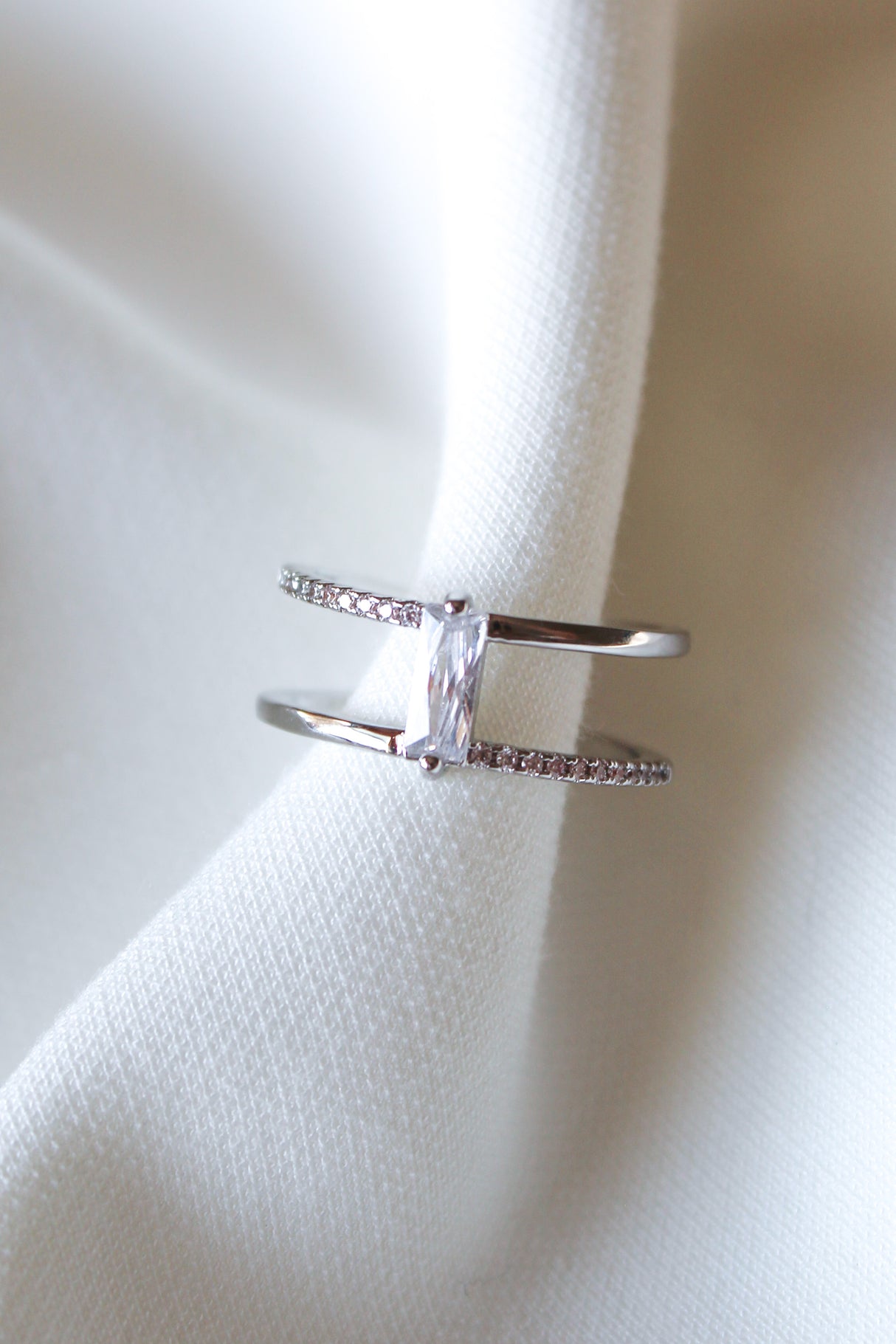 The Cora Ring by Kinsey Designs is an elegant open ring with a rectangular baguette CZ stone set between two bands adorned with small crystals, shown on white fabric.
