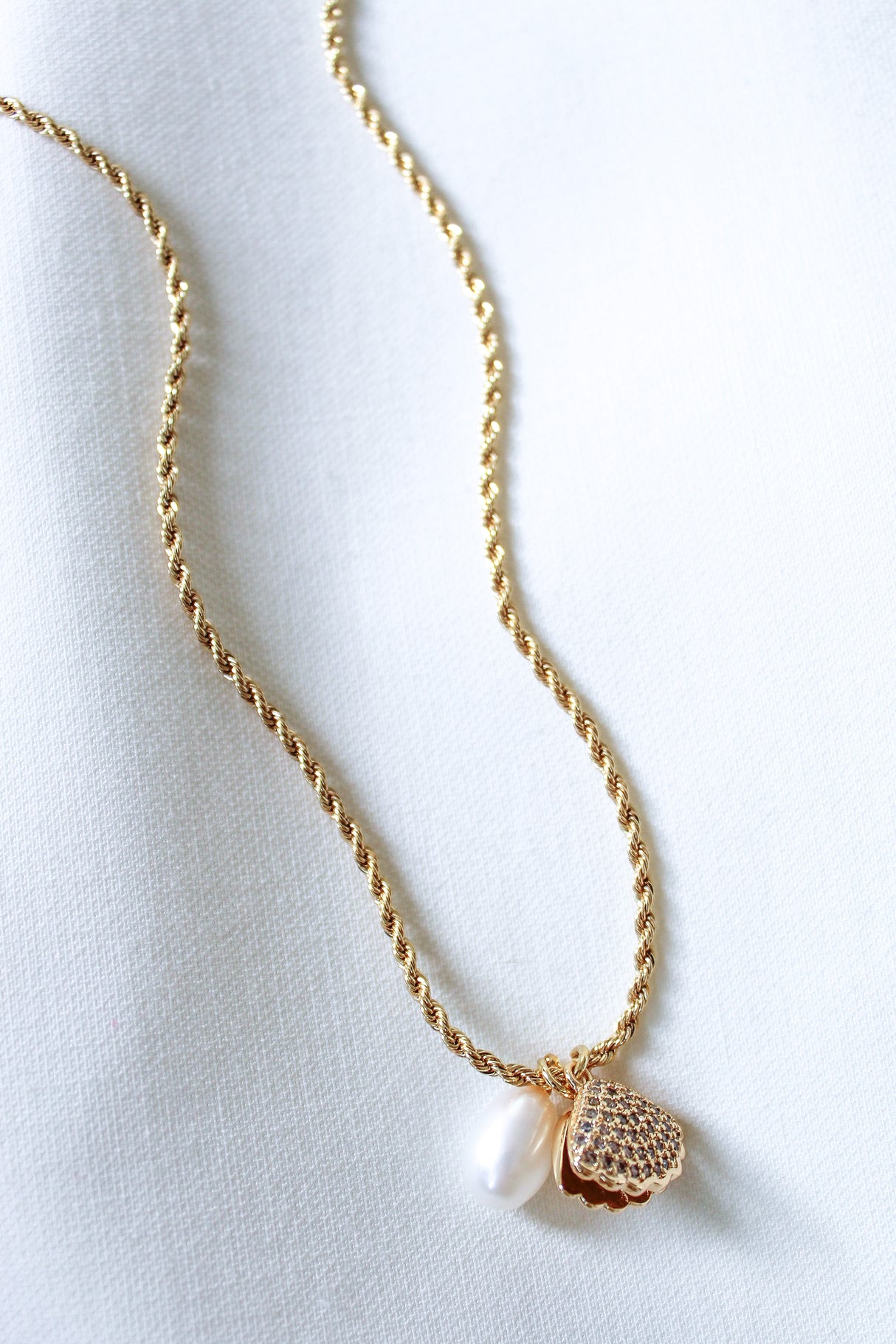 The Cove Pearl Necklace by Kinsey Designs Archives features a white pearl and textured gold heart pendant, elegantly displayed on a white fabric background.