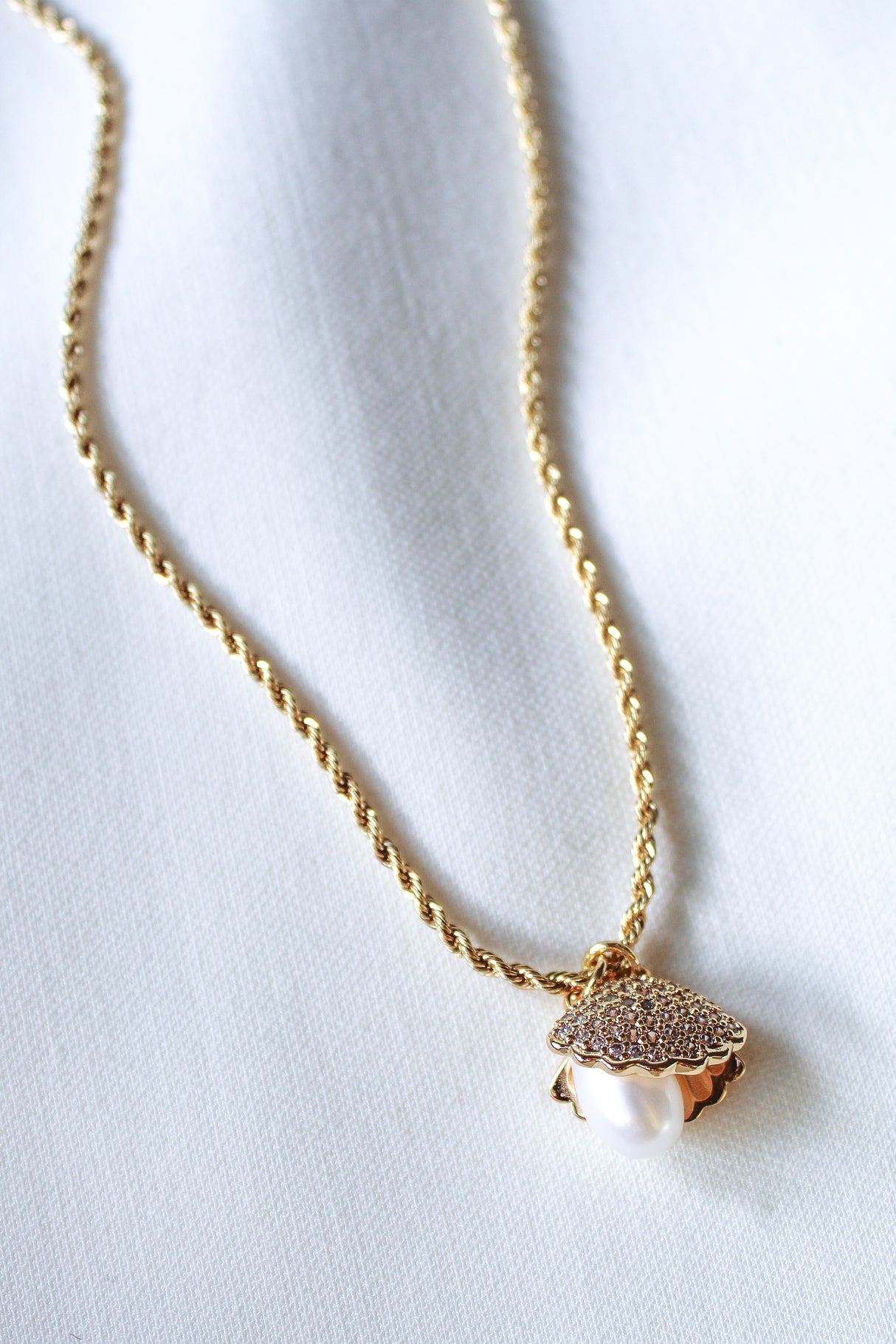 The Cove Pearl Necklace by Kinsey Designs Archives features a gold chain and a seashell locket pendant with a white pearl, elegantly displayed on white fabric.