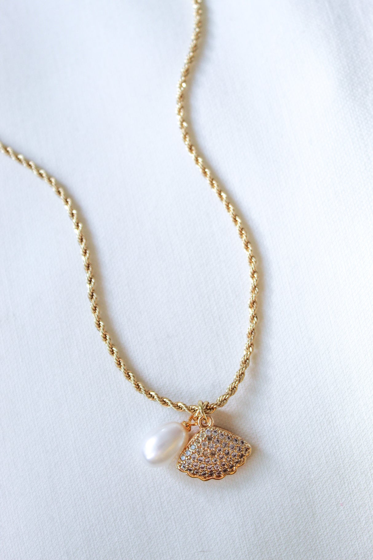 The Cove Pearl Necklace by Kinsey Designs Archives features a gold rope chain with a pearl and a rhinestone-studded gold seashell locket charm, shown on a white fabric background.
