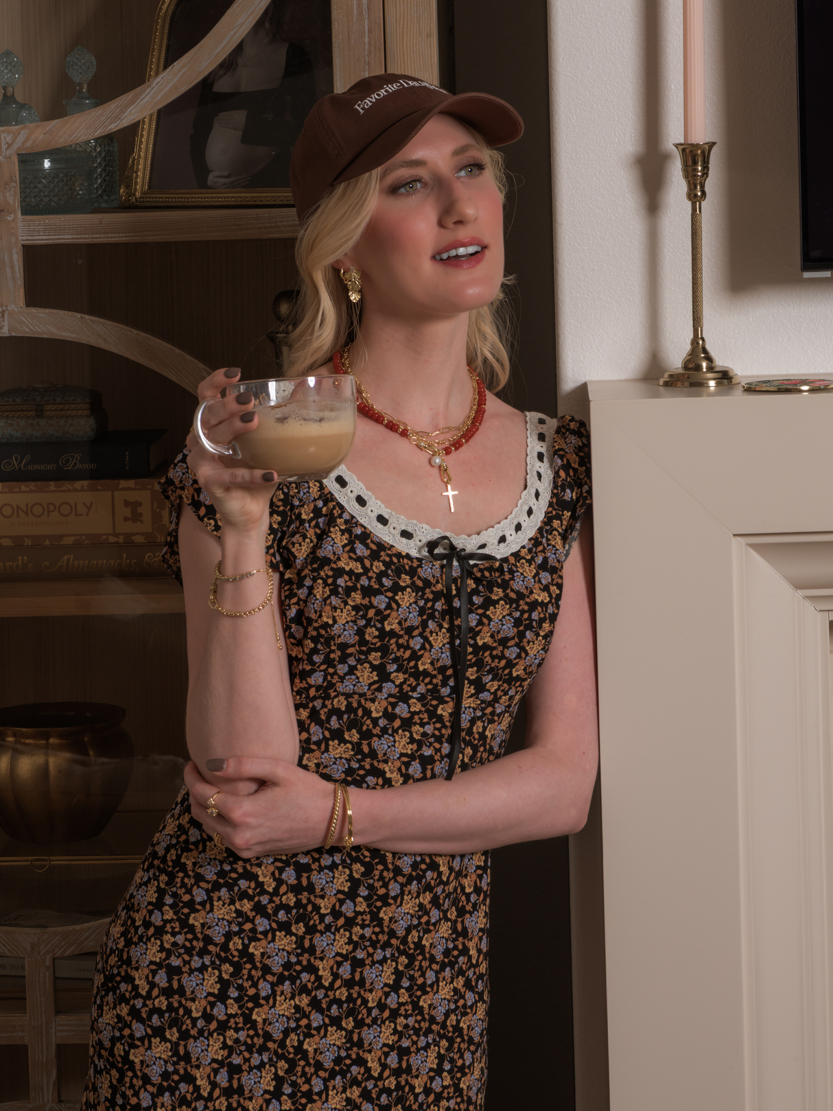 A woman in a floral dress and brown cap holds an iced coffee indoors, smiling as her Kinsey Designs Drew Drop Fan Earring catches the light.