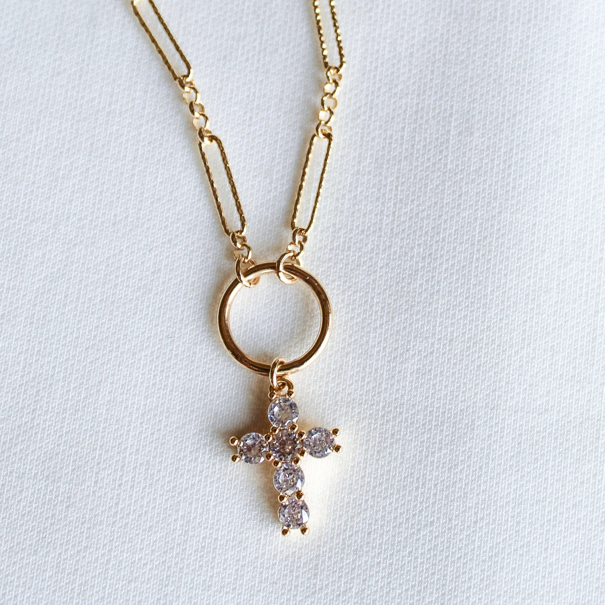 The Denise Necklace by Kinsey Designs features a gold-filled chain and a circular pendant with a crystal-studded cross, shown on a white textured background—an elegant choice for a baptism gift.