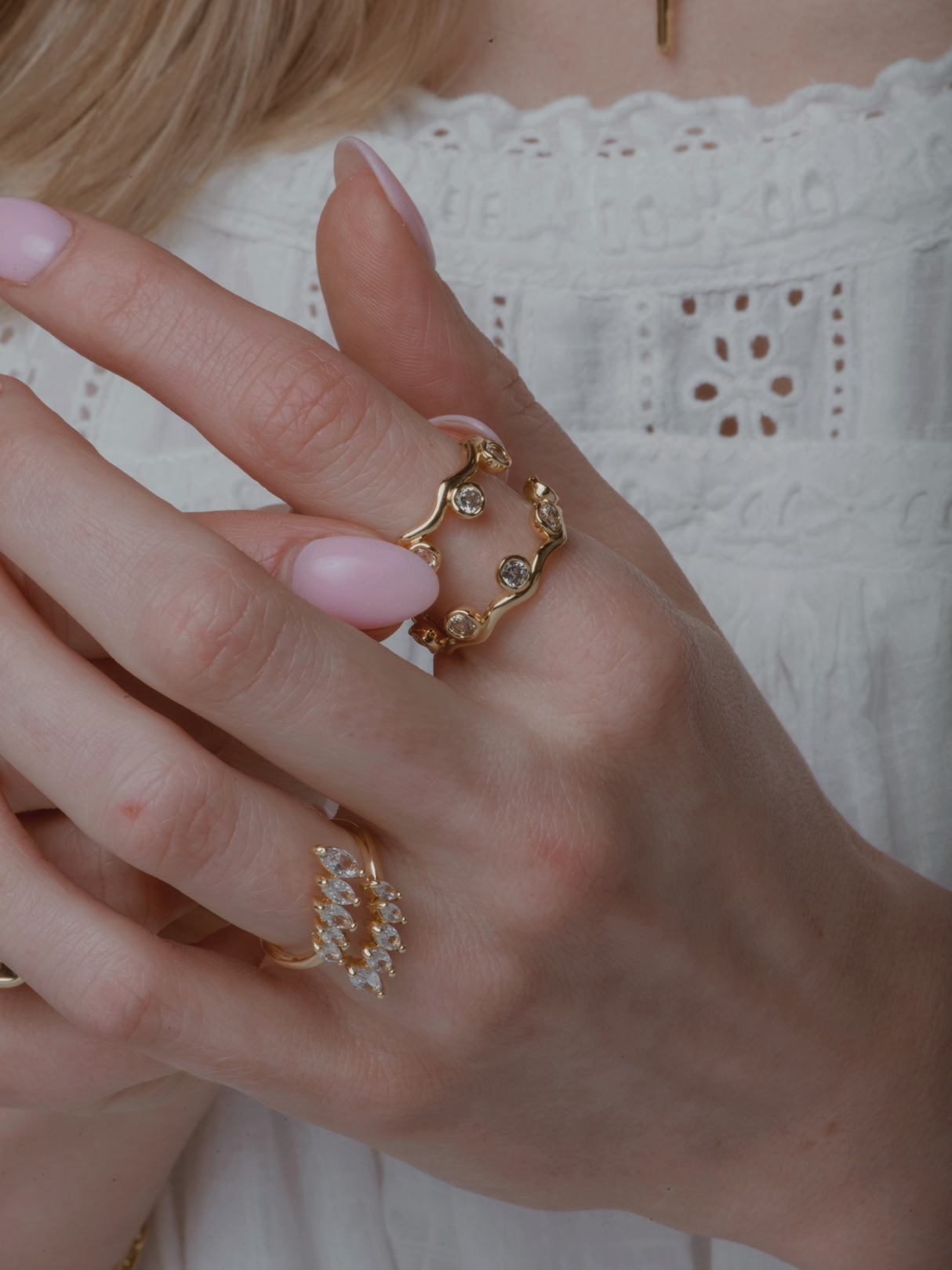 A woman's hand with pink nails showcases the Dove Stacking Ring Set by Kinsey Designs, featuring gemstone-adorned gold circle rings, against a white lace top.