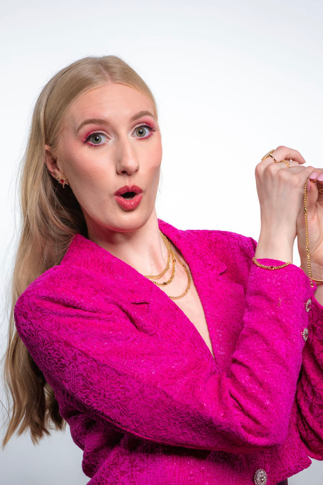 A surprised woman in a bright pink blazer showcases the Kinsey Designs Dublin Bracelet (3 in 1), featuring gold-filled chain layers and sparkling cubic zirconia, against a white background.