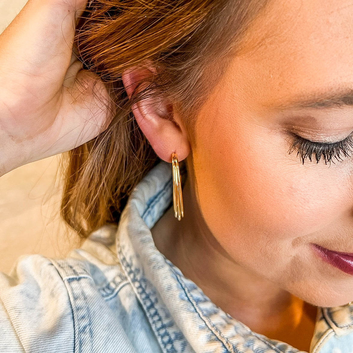 Close-up of a woman wearing Kinsey Designs Archives Dutton Earrings, gold plated hoops measuring 1.5" long and 0.6" wide, with a light denim jacket, touching her hair.
